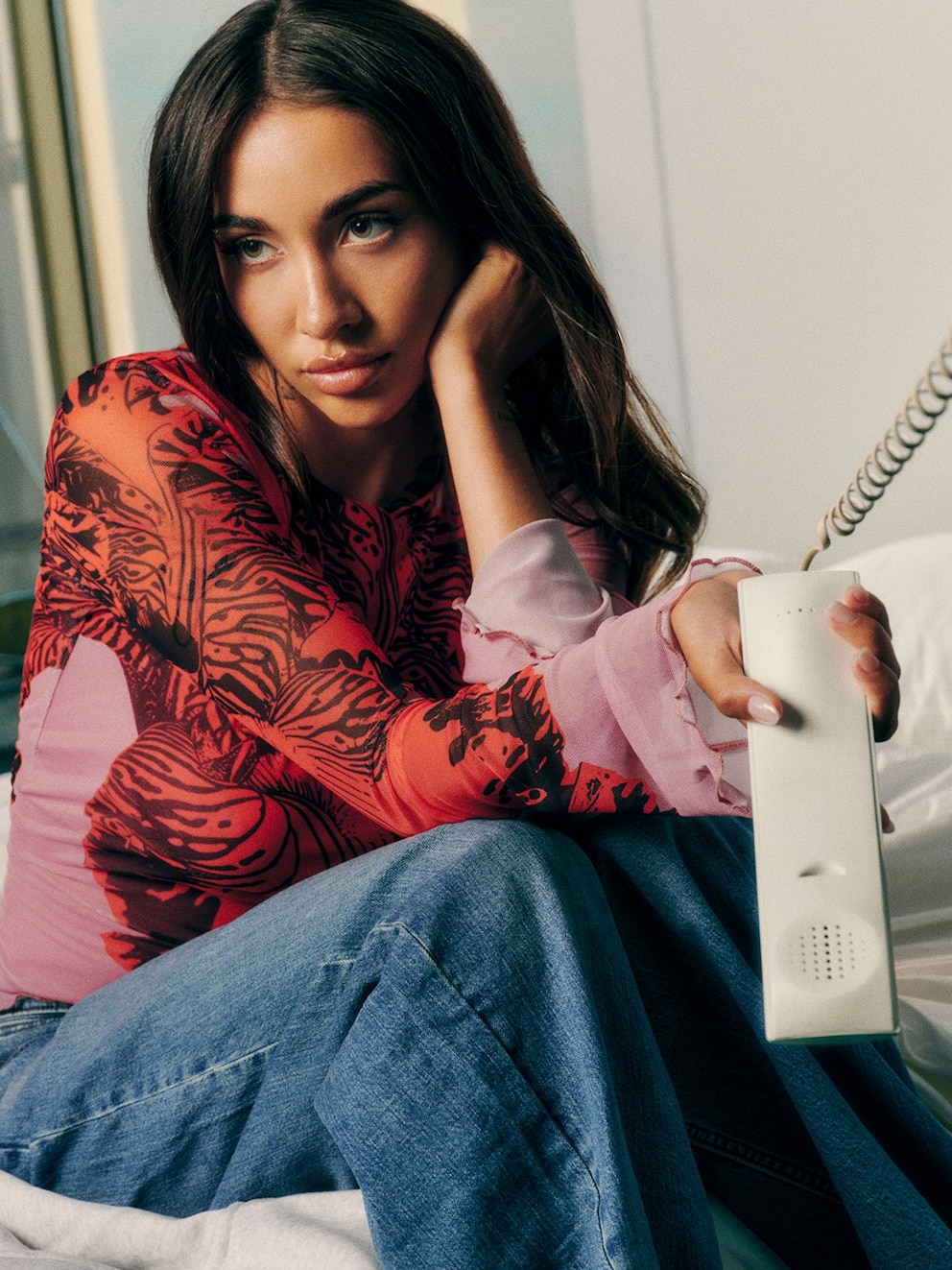 María Becerra posando en una cama con un teléfono en la mano, luciendo una blusa colorida y jeans